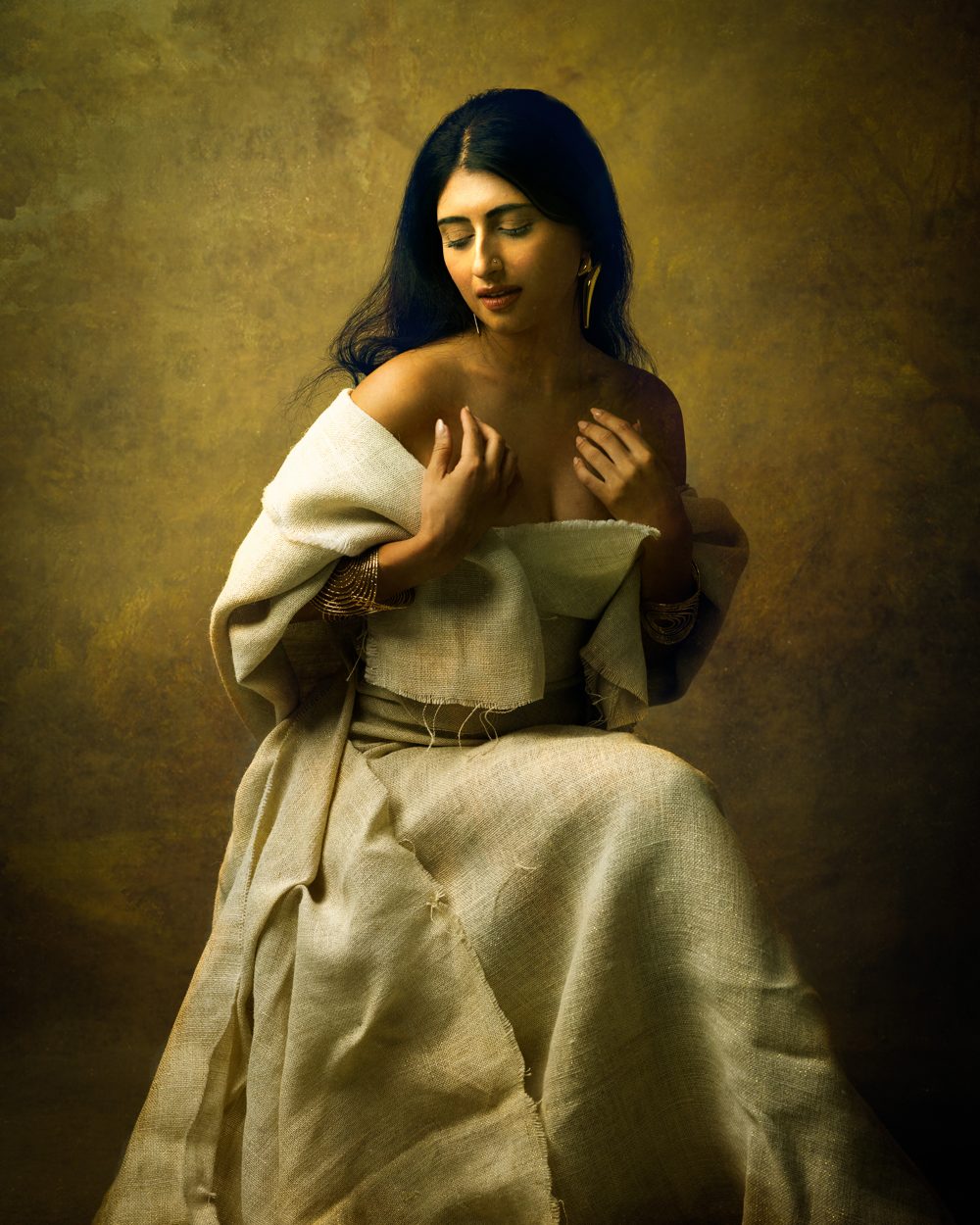 Woman with long dark hair draped in cream linen against a painterly golden backdrop