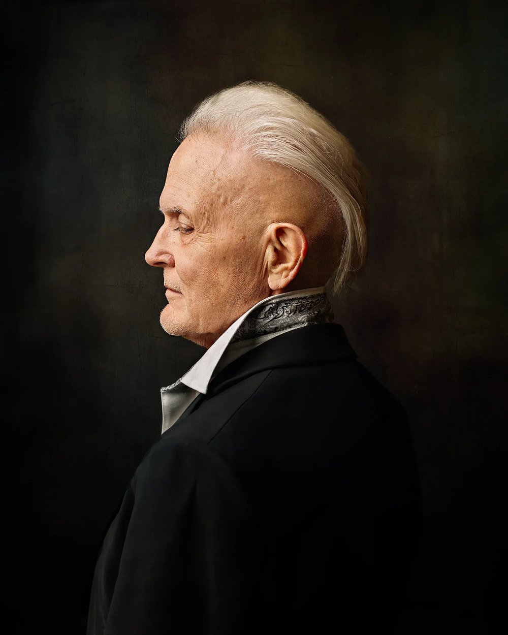 Older man in profile wearing a black suit against a dark background